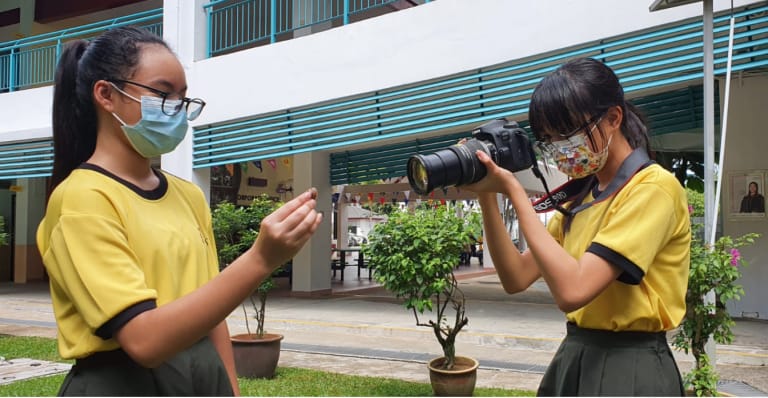 Two masked girls in yellow uniforms outdoors, one photographing a small object held by the other using a DSLR camera.